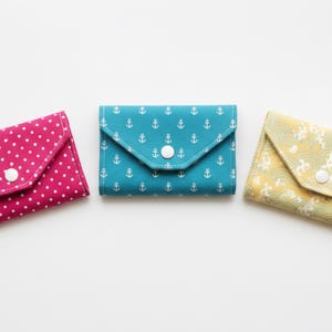 May include: Three rectangular fabric pouches with envelope-style flaps and white snap closures. The left pouch is magenta with white polka dots, the center is teal with white anchor patterns, and the right is yellow with a white wave pattern.