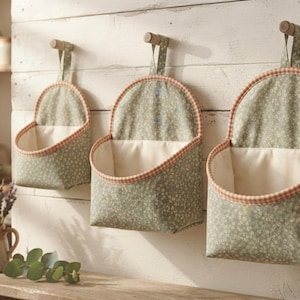 May include: Three hanging fabric wall pockets in varying sizes, featuring a sage green floral print exterior, a solid cream-colored interior lining, and a decorative orange and white checkered trim along the top edge of each open storage pouch.