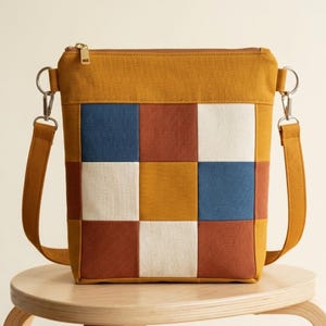 Puede incluir: Un bolso bandolera de color amarillo mostaza con un diseño de patchwork de paneles de tela cuadrados en azul marino, terracota y blanco roto. El bolso tiene un cierre de cremallera superior, una correa a juego y herrajes de metal plateado.