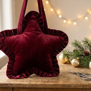May include: A star-shaped tote bag crafted from deep burgundy velvet fabric. The bag features a ruffled velvet trim along its entire perimeter and includes two matching velvet shoulder straps attached to the top points of the star shape.