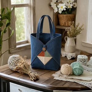 Puede incluir: Un bolso tote de mezclilla azul oscuro con dos asas resistentes y un detalle central de patchwork en forma de diamante en tonos apagados de óxido, verde salvia y crema. El bolso se asegura con un botón de madera redondo en el centro superior.
