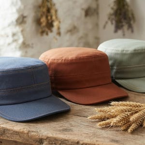 May include: Three cadet caps in blue, rust orange, and sage green. Each cap features a flat top, a short stiff brim, and visible topstitching detail along the crown and brim. The caps are arranged side by side on a wooden surface.