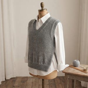 May include: A grey knitted sweater vest with a V-neckline and ribbed trim, layered over a crisp white long-sleeved button-down shirt. The vest features open sides and a straight hemline, displayed on a mannequin torso to show the layered garment.