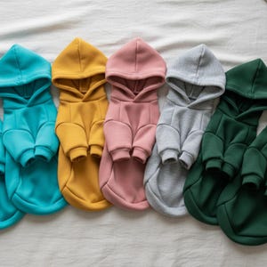 May include: A collection of dog hoodies in various colors, including teal, mustard yellow, dusty rose, gray, and forest green. Each hoodie has a hood and sleeves, designed for a comfortable fit. The hoodies are arranged in a row on a white surface.