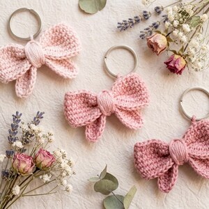 May include: Three crochet bow keychains in varying shades of pink, each featuring a silver metal split ring. The bows are crafted with a textured yarn stitch and a central wrapped knot detail, arranged alongside sprigs of dried lavender and flowers.