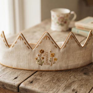 May include: A beige fabric crown featuring a pointed top edge with decorative brown stitching. The center of the crown displays an embroidered number one surrounded by small yellow and white floral motifs with green stems and leaves.