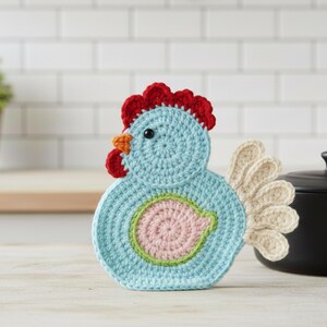 May include: A light blue crocheted chicken ornament featuring a bright red comb and wattle, an orange beak, a small black eye, a pale pink circular wing detail with a light green border, and a cream-coloured fan-shaped tail made of individual petals.