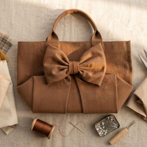 May include: A brown fabric tote bag featuring a large, centered bow on the front panel and two top handles. The bag is surrounded by sewing supplies including fabric scraps, a spool of brown thread, a needle, a pin tin, and metal scissors.