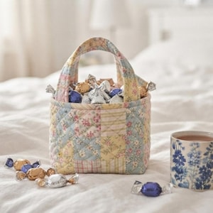 May include: A quilted fabric basket with a patchwork floral pattern in pastel yellow, pink, and blue, filled with foil-wrapped candies. Beside it sits a ceramic mug featuring a blue and white floral print and a small spool of brown twine.