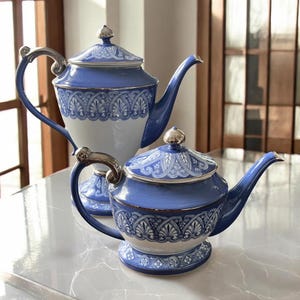 May include: Two blue and white porcelain teapots with silver accents. The teapots have ornate, detailed patterns and matching lids. One teapot is taller than the other. The handles and lids have silver-colored metal accents. The teapots are on a marble-like surface.