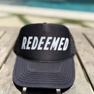 Puede incluir: Gorra de camionero negra con la palabra "REDEEMED" en letras blancas en la parte delantera. La gorra tiene una parte trasera de malla y una visera curva. La gorra está sobre una superficie de madera.