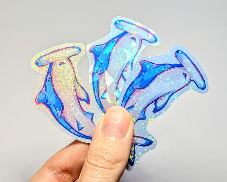 Peut inclure: Trois autocollants holographiques iris&eacute;s repr&eacute;sentant des requins marteaux dans diff&eacute;rentes poses. Les autocollants sont sur un fond blanc et ont un sch&eacute;ma de couleurs bleu et blanc.