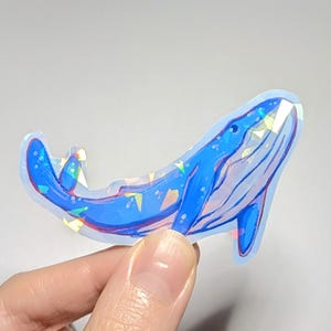 Peut inclure: Un autocollant de baleine bleue avec des accents de paillettes iris&eacute;es. La baleine est tourn&eacute;e vers le haut, la bouche ouverte. L'autocollant est sur un fond blanc.