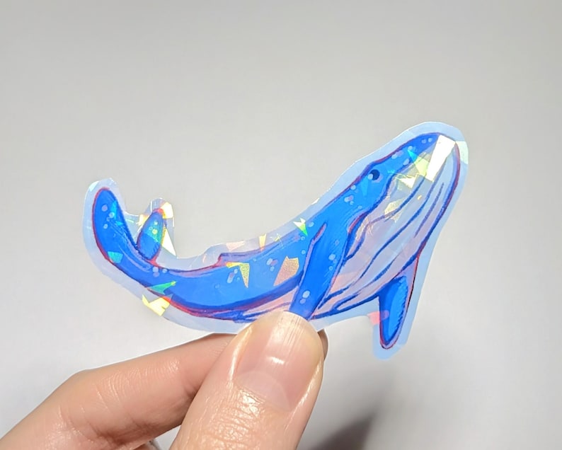 Peut inclure: Un autocollant de baleine bleue avec des accents de paillettes iris&eacute;es. La baleine est tourn&eacute;e vers le haut, la bouche ouverte. L'autocollant est sur un fond blanc.