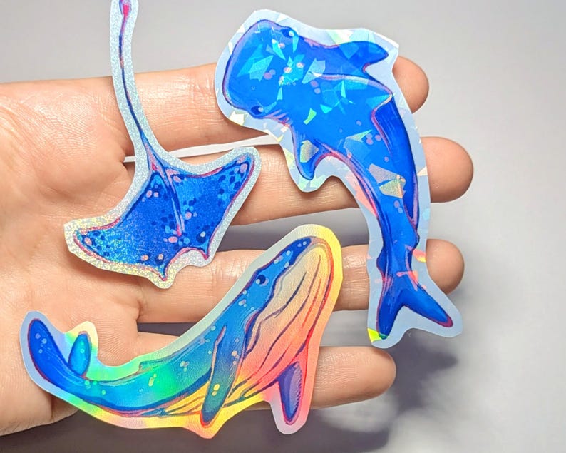 Peut inclure: Trois autocollants holographiques iris&eacute;s de cr&eacute;atures marines&nbsp;: une baleine bleue, une raie manta et un requin. Les autocollants sont sur une main, et la baleine est au premier plan.