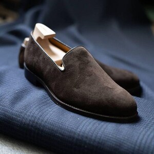 May include: A pair of brown suede loafers with a black sole and a wooden shoe tree inside. The shoes are set on a blue patterned fabric, showcasing the texture and detail of the footwear.
