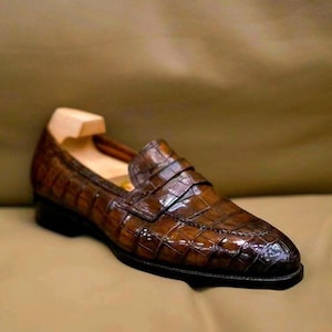 Puede incluir: Un par de mocasines de cuero marr&oacute;n con estampado de cocodrilo. Los mocasines tienen una punta puntiaguda y una suela de cuero.