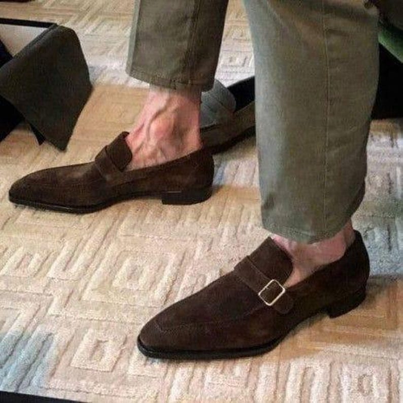 May include: Brown suede loafers with a strap and buckle closure. The shoes are worn with olive green pants. The loafers have a classic design and are suitable for formal or casual wear.