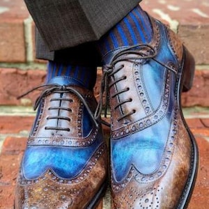 May include: A pair of two-tone leather dress shoes with blue and brown hues. The shoes feature intricate brogue detailing and are laced up. The image also shows blue and brown striped socks and dark brown trousers.