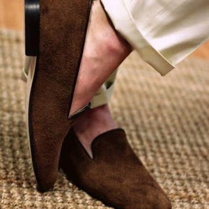 May include: Brown suede loafers with a low heel and a classic slip-on design. The shoes have a dark brown sole and a V-shaped detail on the upper. The loafers are paired with beige trousers.