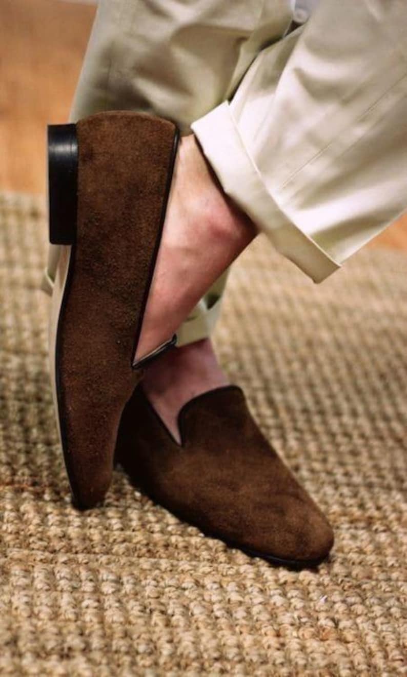 May include: Brown suede loafers with a low heel and a classic slip-on design. The shoes have a dark brown sole and a V-shaped detail on the upper. The loafers are paired with beige trousers.