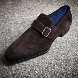 May include: A single brown suede monk strap shoe with a silver buckle. The shoe has a stitched design around the edges and a dark sole. The interior is lined with blue fabric. The shoe is set against a gray background.