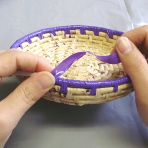 May include: A small, round woven basket with a purple rim and accents. The basket is made of natural-colored straw and is being worked on by a person. The rim is a solid purple color, and the accents are also purple.
