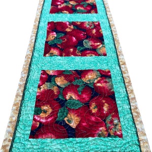 May include: A quilted table runner with a green background and three red apple squares. The runner has a gold border with a small floral pattern.