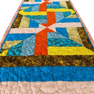 May include: A colorful quilted table runner with geometric patterns in shades of blue, orange, yellow, and brown. The runner has a pink border and is quilted with a decorative stitch.