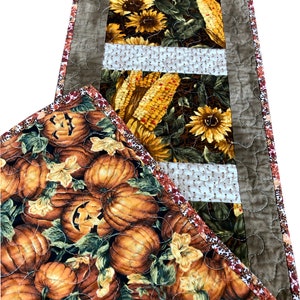 Könnte beinhalten: Ein brauner gesteppter Tischläufer mit Herbstthema. Der Läufer hat drei Paneele mit verschiedenen Designs: Sonnenblumen, Mais und Kürbisse. Der Läufer hat einen braunen Rand mit einem gesteppten Muster.