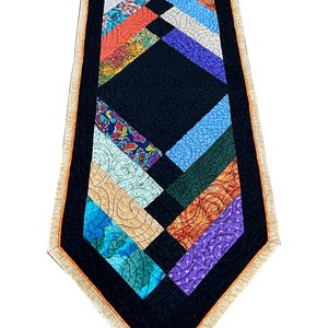 May include: A quilted table runner featuring a black border and a patchwork of colourful fabrics. The design incorporates diagonal stripes in shades of blue, orange, purple, and green, with a central black diamond. The edges are finished with a tan trim.