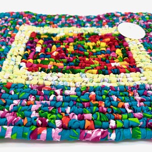 May include: A square, handmade rag rug with a vibrant, colorful design. The rug features a patchwork of fabric strips in shades of blue, pink, green, yellow, red, and purple. The rug's dimensions are approximately 18 inches by 18 inches.