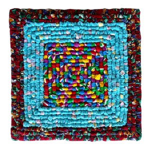 May include: A colorful, square rug made from recycled fabric scraps. The rug has a blue center with a multi-colored square in the middle. The rug is surrounded by a red, white, and blue border.