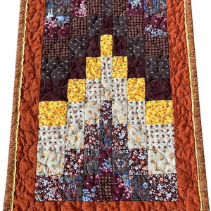 May include: A quilted wall hanging with a geometric pattern in shades of brown, yellow, and white. The quilt is framed with a brown and yellow border.