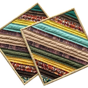 May include: Two quilted placemats with a striped pattern in shades of brown, yellow, green, and teal. The placemats have a cream-colored border.