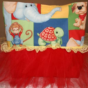 May include: A colorful fabric box featuring cartoon animals: an elephant, monkey, turtle, and dog. The box is trimmed with yellow lace and has a red tulle skirt, creating a whimsical, playful design. Ideal for a child's room.