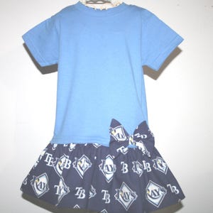May include: A light blue t-shirt dress with a navy blue skirt. The skirt features a repeating pattern of white diamonds with the letters "TB" and "RAYS" inside. A bow detail is on the front.