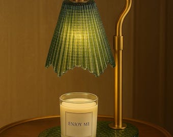 May include: A green glass lamp with a brass-colored curved arm and base. The lamp has a pleated shade and sits above a candle in a glass jar. The candle has a label that reads "ENJOY ME". The lamp and candle sit on a small, round table.