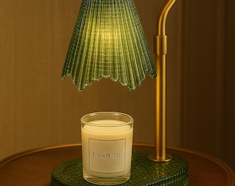 May include: A decorative table lamp with a green, fluted lampshade and a gold-colored metal frame. A candle in a glass jar with the text "ENJOY ME" sits on a green, fluted base. The lamp is on a wooden table.