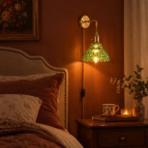 May include: A wall-mounted lamp featuring a brass-toned metal fixture and a scalloped, translucent green glass shade. The lamp includes a braided cord with an integrated switch, emitting a warm glow from the light bulb inside.