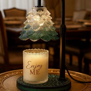 May include: A decorative lamp with a green and clear glass shade shaped like a Christmas tree. A candle with the words "Enjoy ME" is on a green base. The lamp has a black metal frame and sits on a gold-colored table.