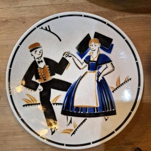 May include: A white ceramic plate with a black border and a decorative illustration of a dancing couple in traditional attire. The man wears a black suit and the woman a blue dress. The plate has a cracked glaze.
