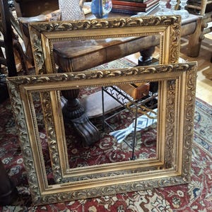 May include: Two ornate, gold-colored picture frames with detailed scrollwork. The frames are rectangular and empty, resting on a patterned red rug. The frames have a vintage, antique aesthetic.