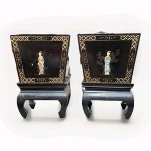 May include: Pair of black wooden chairs with ornate detailing. Each chair features a square back with a decorative border and a central image of a person. The chairs have curved legs and are set against a white background.