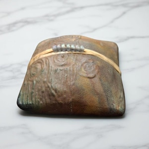 May include: A handcrafted, rectangular ceramic dish with a textured, mottled brown and green surface. A thin, light-colored band with small, gray beads is wrapped around the center. The dish has a unique, organic shape.