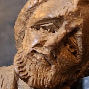 May include: A close-up of a carved wooden sculpture of a man with a beard. The sculpture shows detailed lines and textures, highlighting the artist's skill. The man's facial features are prominent, with emphasis on the eyes and beard. The wood has a warm, natural colour.