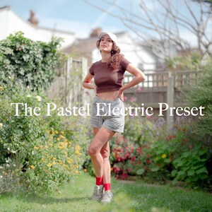 May include: A woman wearing a brown crop top and black and white checked shorts stands in a garden. She is wearing a white cap and red socks. The garden has green grass and colourful flowers.