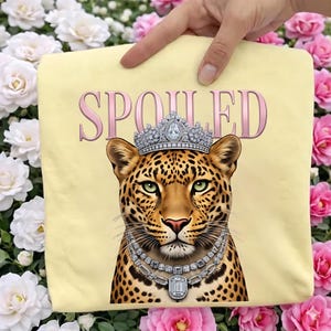 May include: A light yellow square pillow with the word "SPOILED" in pink script. The pillow features a regal leopard wearing a jeweled crown and necklace. The leopard has green eyes and a spotted coat.