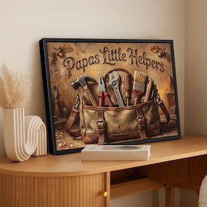 May include: Framed artwork featuring a canvas tool bag filled with tools, including a hammer, pliers, and a wrench. The text "Papas Little Helpers" is displayed above the bag. The artwork is set against a wooden background.