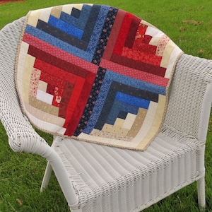 May include: A red, white, and blue log cabin quilt with a white wicker chair in the background.